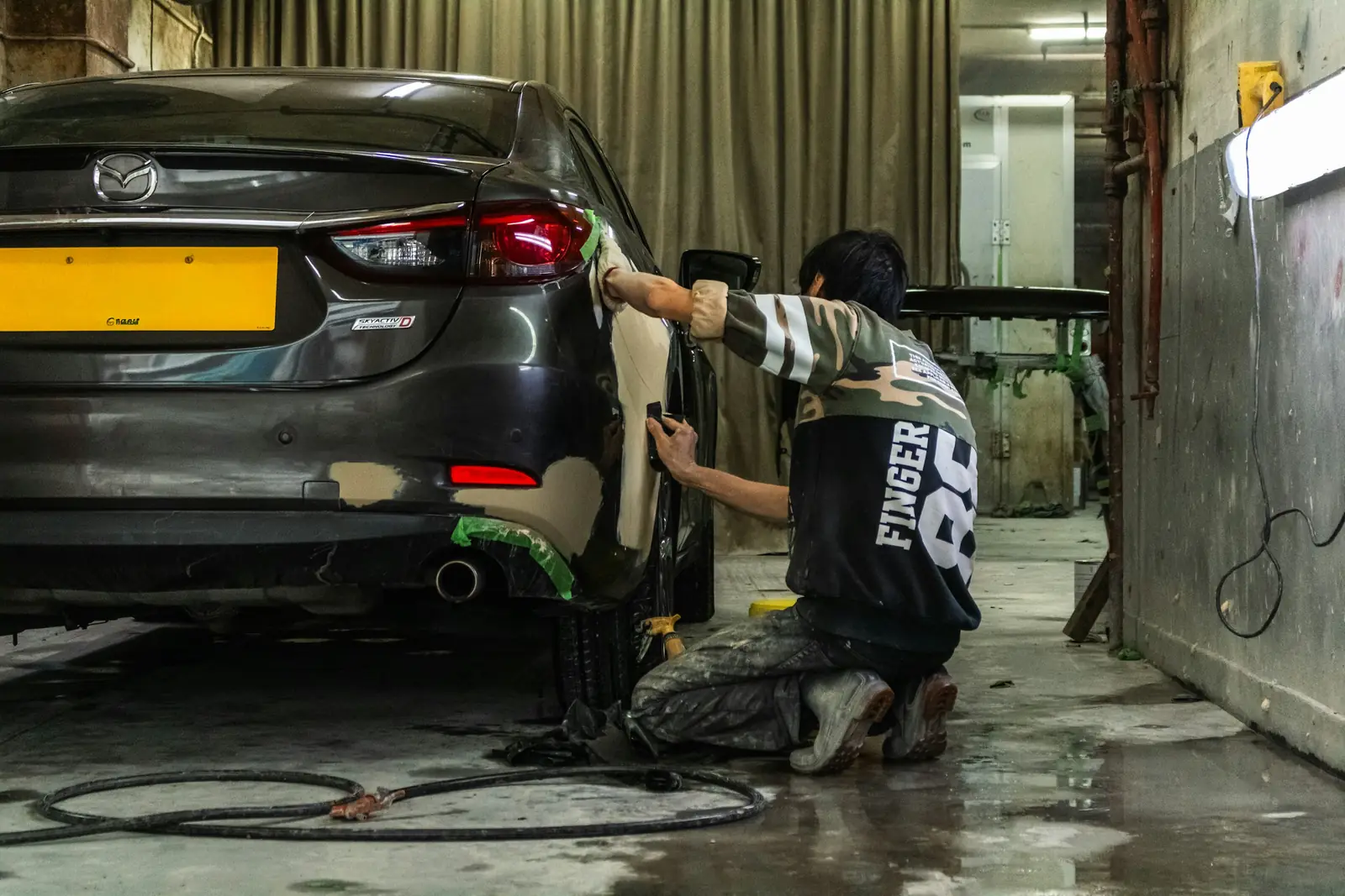 Repairer kneeling at a rear quarter panel, sanding filler back to shape
